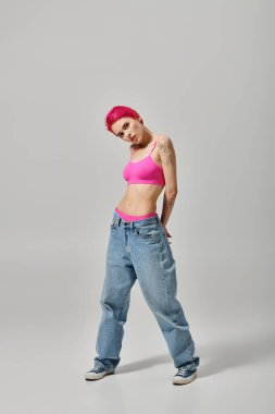 Pembe bluzlu ve kot pantolonlu çekici genç bir kadın gri arka planda kameraya bakıyor, moda.