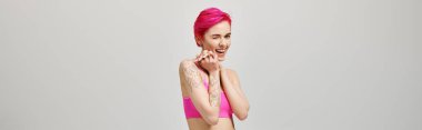 excited young woman with pink short hair posing in crop top and winking on grey backdrop, banner clipart