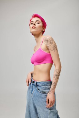 Pembe kısa saçlı kendine güvenen bir kadın. Kırpılmış bluzla poz veriyor ve gri arka planda kot pantolon giyiyor.