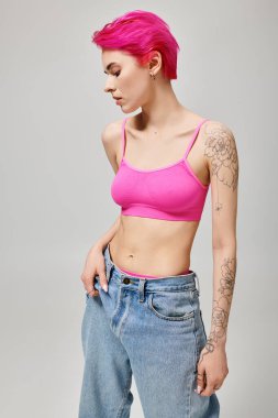 confident and tattooed young woman with pink hair posing in crop top and jeans on grey background clipart