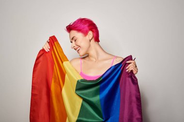 Memnuniyet portresi ve pembe saçlı genç bir kadın gri arkaplanda lgbt gökkuşağı bayrağıyla poz veriyor.