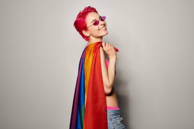 Genç ve memnun kadın aktivist pembe saçlı ve güneş gözlüklü lgbt gökkuşağı bayrağıyla poz veriyor.