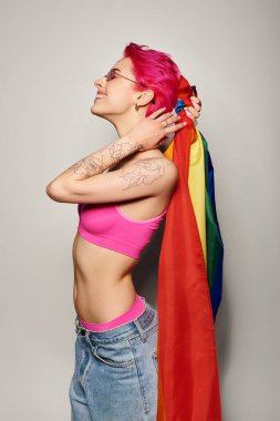 Pembe saçlı ve güneş gözlüklü genç ve neşeli kadın gri arka planda lgbt gökkuşağı bayrağıyla poz veriyor.