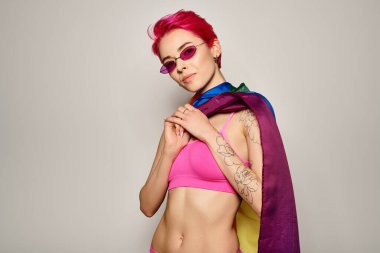 Pembe saçlı ve şık güneş gözlüklü genç bir kadın gri arka planda lgbt gökkuşağı bayrağıyla poz veriyor.