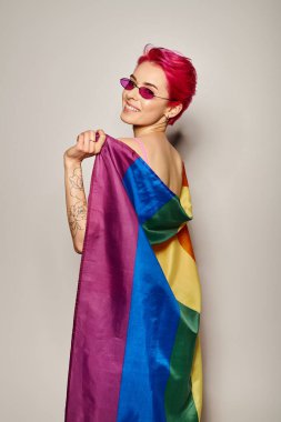 young happy female activist with pink hair and sunglasses posing with lgbt rainbow flag on grey clipart