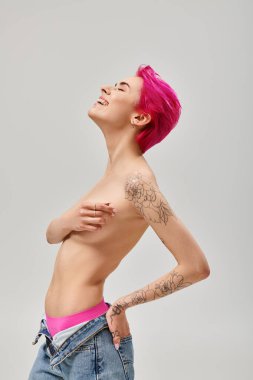 Dövmeli ve üstsüz, pembe saçlı, göğüslerini kapatan ve gri arka planda gülümseyen genç bir kadın.