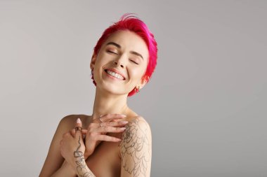Gri arka planda çıplak omuzları ve dövmesi olan neşeli ve üstsüz genç bir kadının portresi.