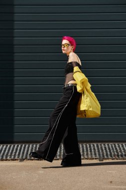 pink haired young woman in yellow sunglasses and stylish attire walking on urban street outdoors clipart
