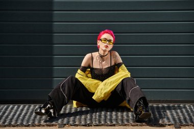 Pembe saçlı, dövmeli, sarı güneş gözlüklü ve şık giyinmiş genç bir kadın.