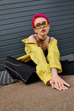 Dövmeli, pembe saçlı, sarı güneş gözlüklü ve açık havada moda sokak kıyafetleri giyen genç bir kadın.