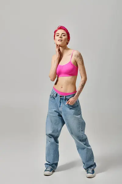 stock image cheerful young woman with pink hair and tattoos posing on grey backdrop with hand in pocket, fashion