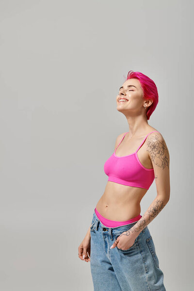 joyful young female model in stylish outfit with tattoos smiling and posing with hand in pocket
