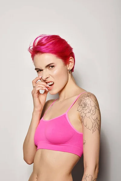 stock image emotional tattooed young woman with pink hair posing in crop top and biting nail on grey backdrop