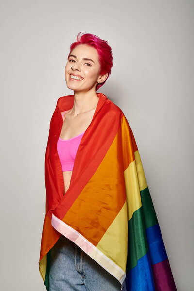portrait of cheerful young woman with pink hair posing with lgbt rainbow flag on grey backdrop