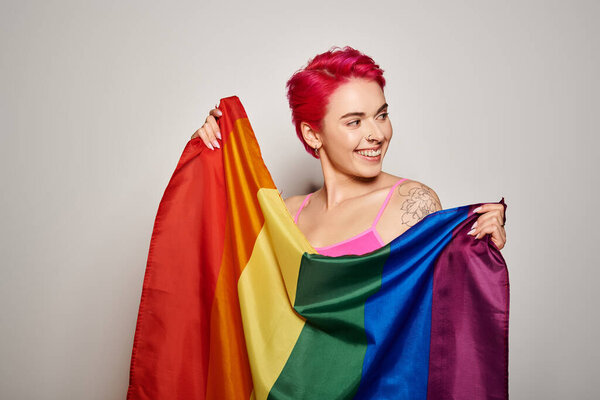 portrait of pleased female activist with pink hair posing with lgbt rainbow flag on grey backdrop