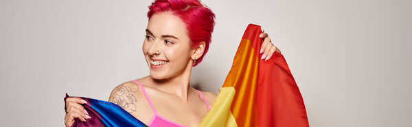 portrait of pleased female activist with pink hair posing with lgbt rainbow flag on grey, banner
