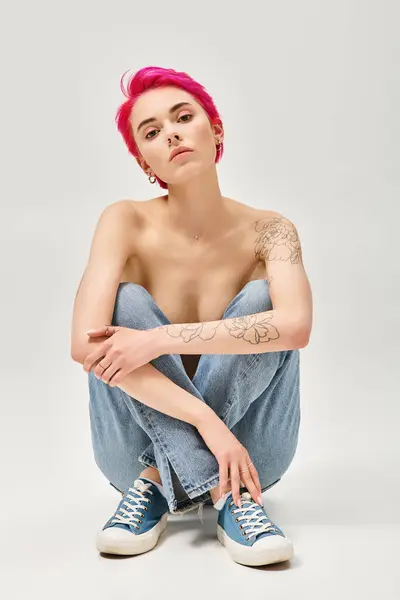 Stock image tattooed and topless woman with pink hair sitting in denim blue jeans on grey background, sexy