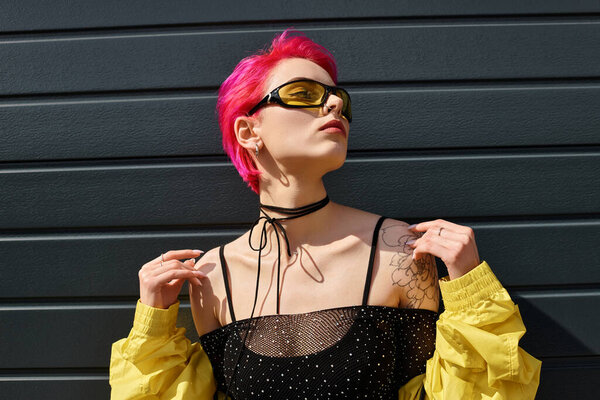 pink haired young woman in yellow sunglasses and stylish outfit posing on urban street outdoors