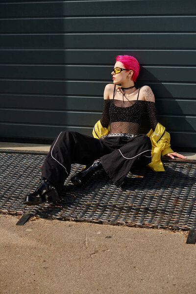 young woman with pink hair and tattoo posing in sunglasses and trendy streetwear on urban street