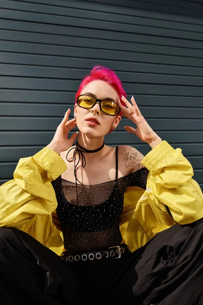 stylish chic woman with pink hair and tattoo posing in sunglasses and trendy streetwear on street