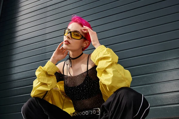 young chic woman with pink hair and tattoo posing in sunglasses and trendy streetwear on street