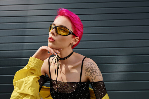 pensive young woman with pink hair and tattoo posing in sunglasses and trendy streetwear on street