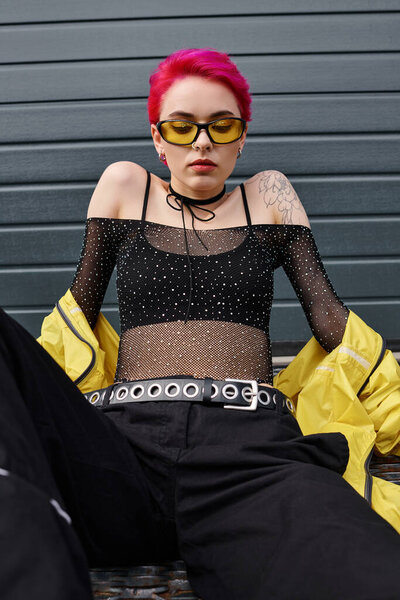 young woman with pink hair and tattoo posing in yellow sunglasses and trendy streetwear on street