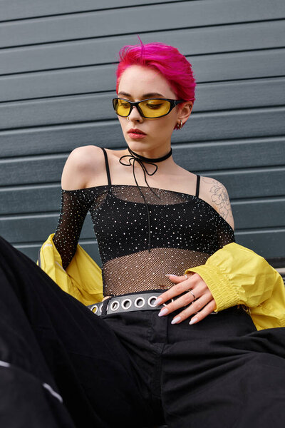 pretty young woman with pink hair and tattoo posing in sunglasses and trendy streetwear on street