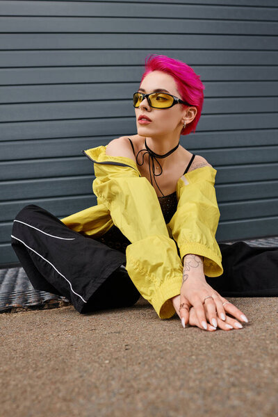 tattooed and pink haired young woman in yellow sunglasses and stylish attire posing on urban street