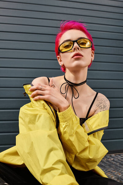 pink haired young woman in trendy sunglasses and stylish attire biting lip and looking at camera