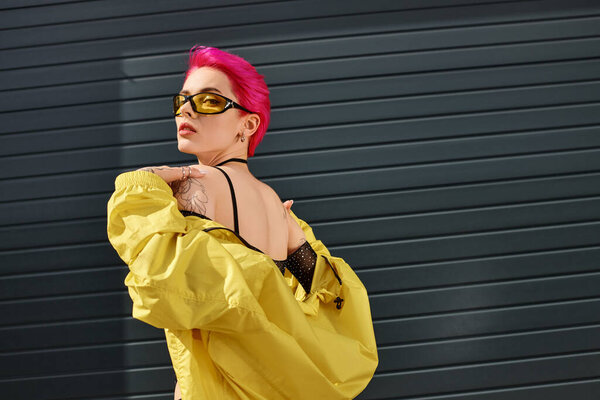 pink haired young woman in yellow sunglasses and trendy outfit posing and looking at camera