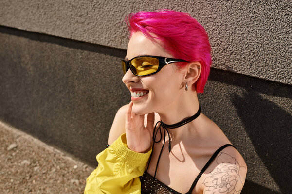 portrait of happy pink haired woman in sunglasses and trendy outfit posing while looking away