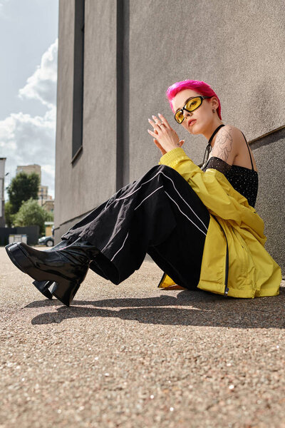 pink haired woman in yellow sunglasses and trendy outfit looking at camera and posing on street