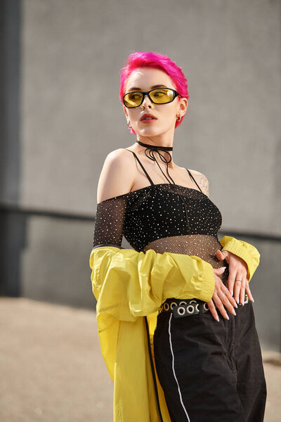 portrait of pink haired young woman in yellow sunglasses and trendy outfit posing on urban street