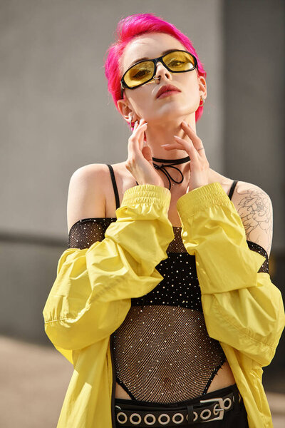 portrait of pink haired young woman in sunglasses and trendy outfit looking at camera outdoors