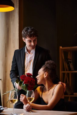 handsome man in suit holding roses near happy african american woman in restaurant on Valentines Day clipart