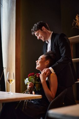 Arkasında duran takım elbiseli adamla el ele tutuşan mutlu Afro-Amerikan kadını, kırmızı güller.