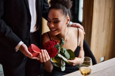 gentleman gifting heart-shaped box to happy african american woman with roses on Valentine`s day clipart