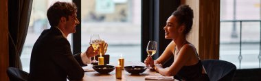 happy interracial couple in elegant attire holding glasses of wine during date in restaurant, banner clipart