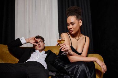 Kıvırcık Afrikalı Amerikalı kadın elinde bir bardak şarapla kadife koltukta takım elbiseli bir adamla oturuyor.