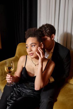 Zarif, genç, Afro-Amerikalı bir kadın elinde bir bardak şarapla kadife koltukta bir adamla oturuyor.