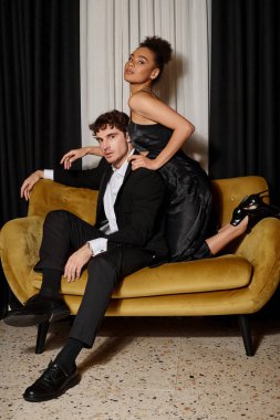 Kıvırcık Afrikalı Amerikalı kadın takım elbiseli yakışıklı adamı kucaklıyor ve kadife koltukta poz veriyor.