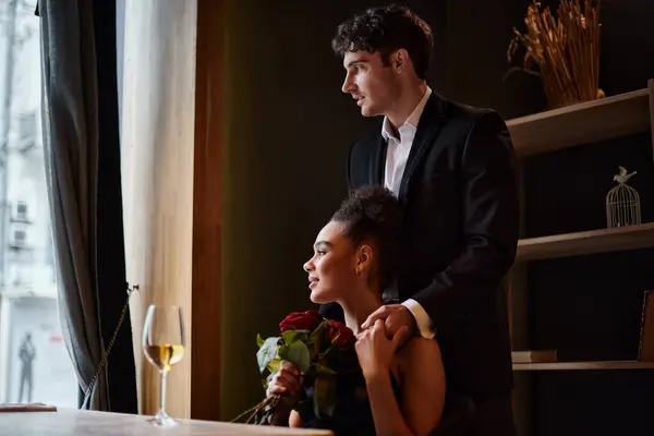 stock image pleased african american woman holding hands with man in suit standing behind her, red roses