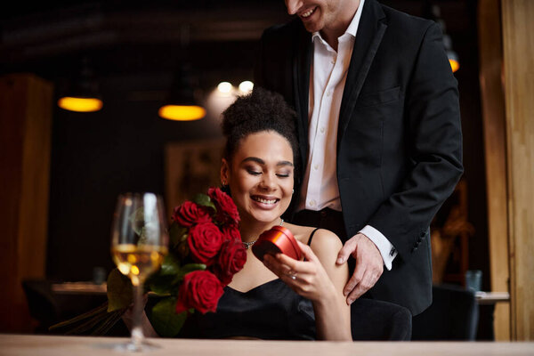 happy man gifting heart-shaped box to cheerful african american woman with red roses on 14 February