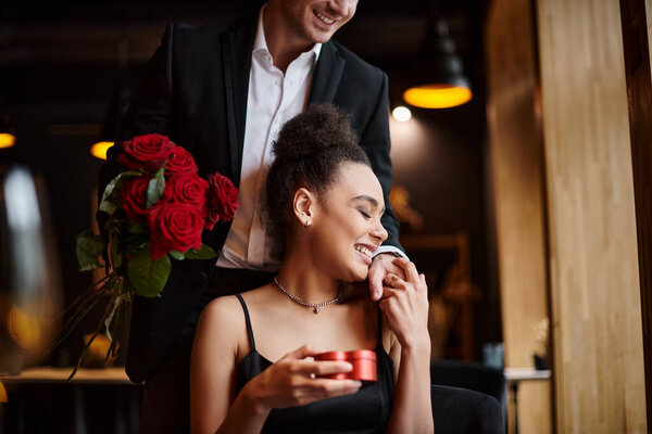 man holding red roses near happy african american woman with heart-shaped box on Valentines day