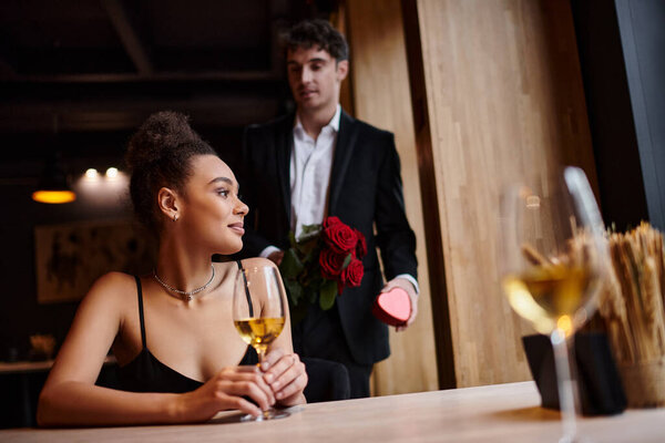 happy man holding red roses and heart-shaped box near african american woman on Valentines day