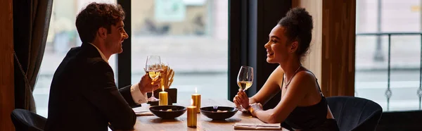 happy interracial couple in elegant attire holding glasses of wine during date in restaurant, banner