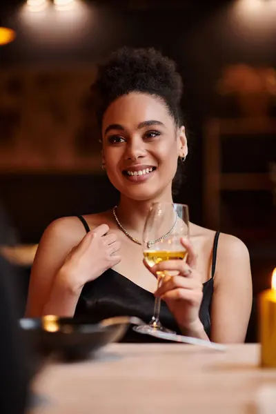 stock image cheerful african american lady smiling and holding glass of wine during date on Valentines day