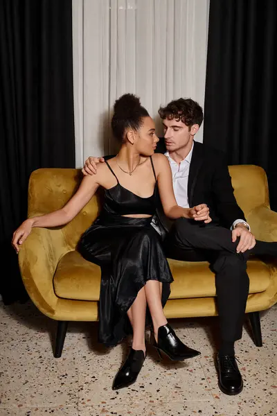 young multicultural couple in elegant attire sitting on velvet sofa, african american woman and man