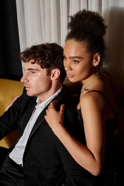 attractive african american woman in dress hugging man in formal wear and posing on velvet sofa